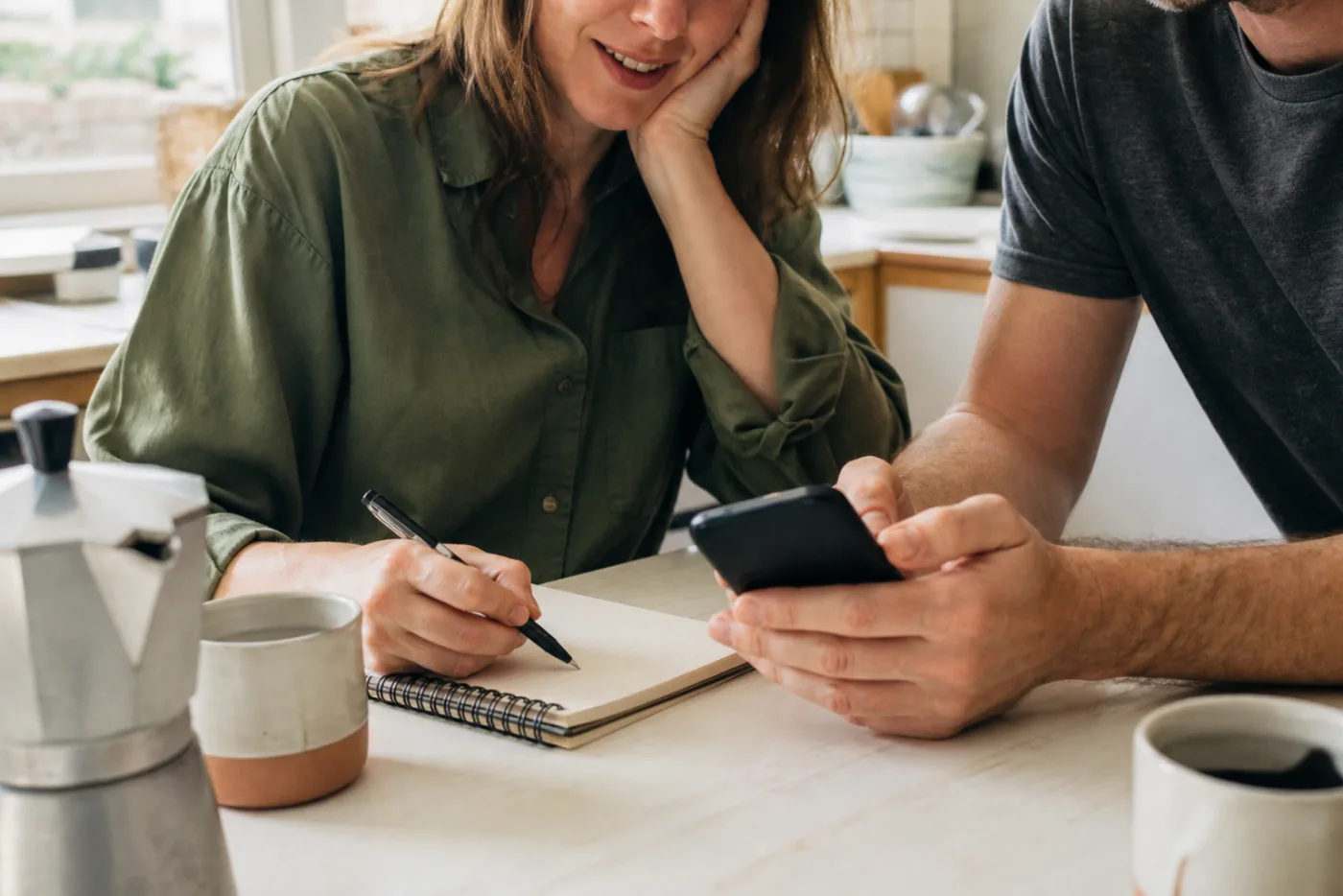 Two neurodivergent adults at a kitchen table, looking at a phone and communication notes together.